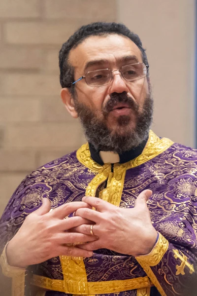 A bearded man with glasses wears purple liturgical vestments with gold trim and embroidery. His hands are clasped at his chest, revealing a wedding band, and he has a thoughtful expression.